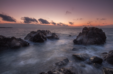 A rugged coastal seascape at sunrise on the Cornish coast. Waves flowing over dark, granite rocks as the sun sets over the horizon in Cornwall.の写真素材