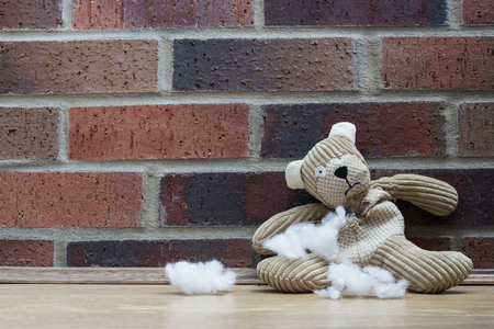 A sad and abandoned teddy bear sitting alone against a brick wall with stuffing and filling falling out of his ripped and torn tummy.の写真素材
