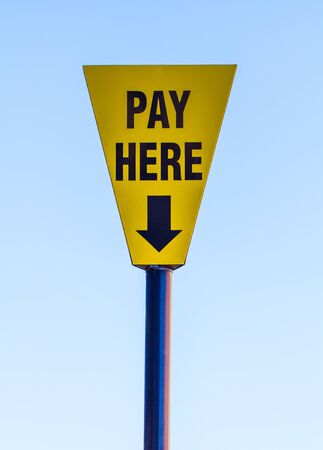 A yellow and blue, triangular sign informing people to Pay Here with an arrow pointing down to a ticket machine with a blue sky background.の写真素材