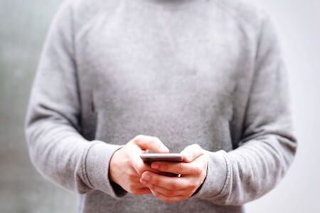 A young man in grey sweatshirt holding his mobile phone whilst texting in a contemporary, midsection...