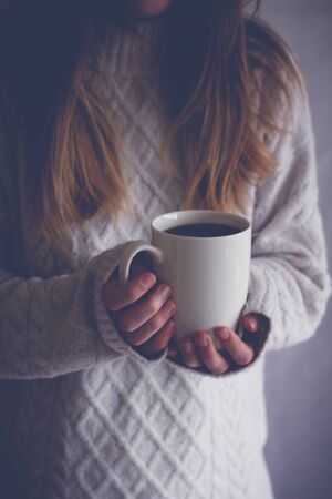 A midsection of a blonde haired girl holding up a white cup indoors and in the shadows.の写真素材
