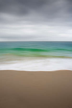An abstract background image of an emerald green ocean with white waves over a sandy beach using a blurred motion panning effect to create a beautiful nature canvasの素材