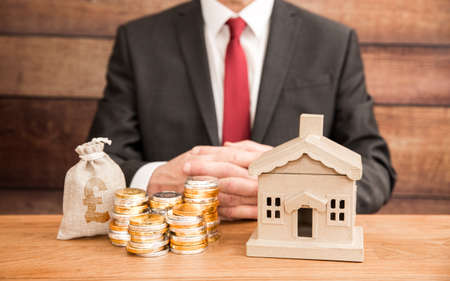 A housing concept of the cost and price of homes with a real estate agent or mortgage broker sitting with a property and money to represent fees, mortgage and house prices.の写真素材