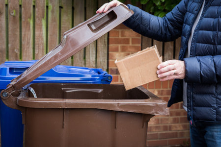 A responsible man placing a brown cardboard box into a kerbside recycling bin ready for collection and recycling by the Council in the UKの写真素材