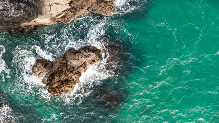 Aerial view directly above white waves breaking on harsh stone rocks in a rugged environment on the...