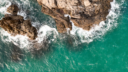 Aerial view directly above white waves breaking on harsh stone rocks in a rugged environment on the Cornwall coast with copy space above emerald green oceanの写真素材