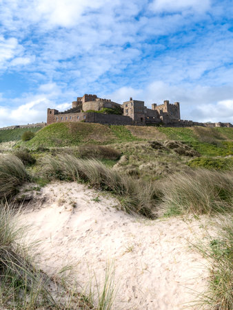 BAMBURGH CASTLE, NORTHUMBERLAND, UK - APRIL 23, 2024.  Vertorama landscape panorama view of Bamburgh castle and sand dunes on the beautiful Northumberland coast with blue sky and sunshineのeditorial素材
