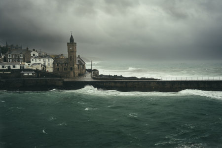 Dark storm clouds with gale force winds and waves crashing against the harbour wall and church in Porthleven on the Lizard peninsula in Cornwallの写真素材