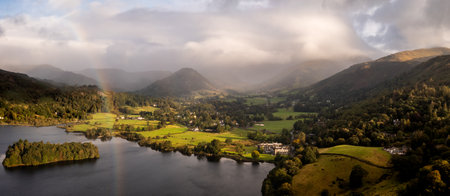 Aerial panoramic landscape of the village and lake of Grasmere in The Lake District National Park with calm water and sunshine in the valley and a brooding storm on the hills creating a rainbowの写真素材