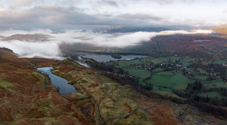 Aerial panorama landscape view of the lakeside village of Grasmere and lake in The English Lake District National Park from Alcock Tarn with surrounding mountains and cloud inversion on a cool Spring morningの写真素材