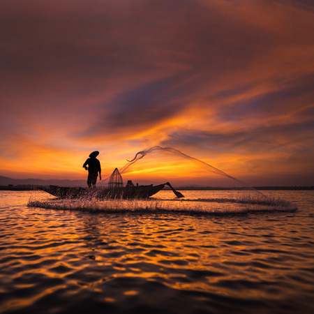 Silhouette of asian fisherman on wooden boat in action casting a net for catching freshwater fish in nature river in the early morning before sunriseの写真素材