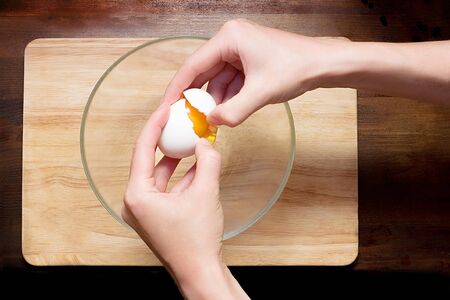 Hands break an egg into a glass bowl on a wooden dark and light table
