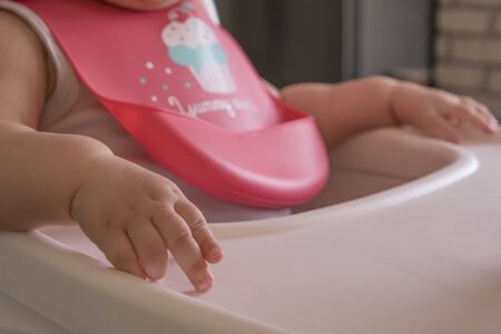 Baby girl hand and fingers in focus while sitting on a feeding chair and a bib onの写真素材