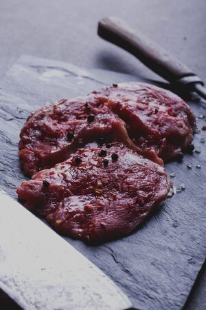 Raw fresh meat Ribeye Steak, with seasoning and meat axe and fork on black slate boardの写真素材