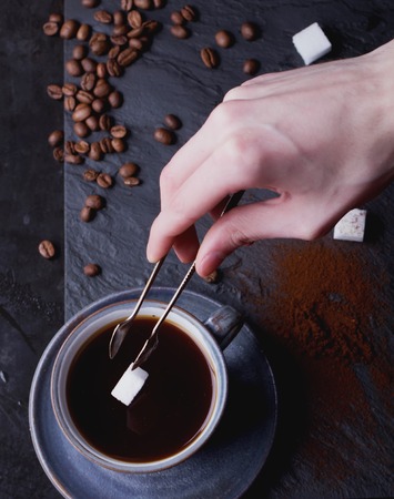 A cup of coffee on black slate board with coffee beans, sugar and tiny sugar tongs. Male hands putting sugar in the cup. Top Viewの写真素材