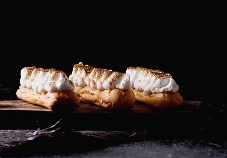 Home made eclairs decorated with white meringue on the rustic wooden serving board black background....