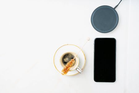 Black smartphone, wireless charger and cup of coffee over white background. Top view. Copy space.の写真素材
