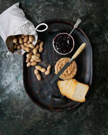 Peanut butter, raspberry jelly and slices of toasted bread on the tray over rustic metal background. Top view. Breakfast sandwiches preparation. Male hand spilling out peanuts on the tray.の写真素材