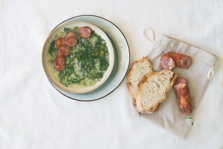 Caldo Verde Soup with greens and chopped chorizo on the top in ceramic bowl over ceramic plate. Pieces of bread and chorizo over cotton bag. Top Viewの写真素材