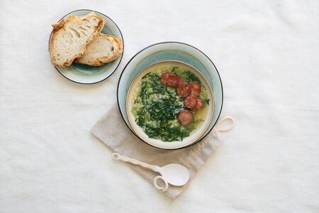 Caldo Verde Soup with greens and chopped chorizo on the top in ceramic bowl over ceramic plate on cotton bag with ceramic spoon. Pieces of bread on the plate. Top Viewの写真素材