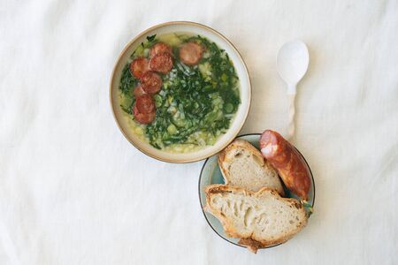 Caldo Verde Soup with greens and chopped chorizo on the top in ceramic bowl with ceramic spoon. Pieces of bread and chorizo on ceramic plate. Top Viewの写真素材