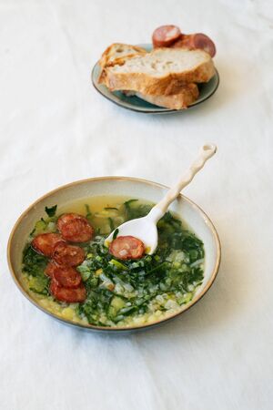 Caldo Verde Soup with greens and chopped chorizo on the top in ceramic bowl with ceramic spoon. Pieces of bread and chorizo on ceramic plateの写真素材