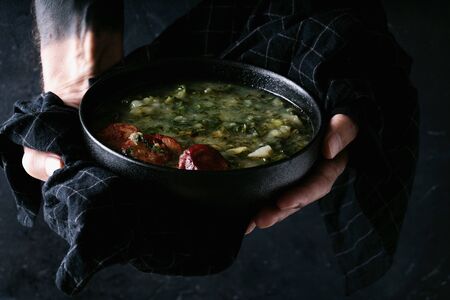 Caldo Verde Soup with greens and chopped chorizo on the top in the bowl in male hands on a dark background. Selective focus. Close upの写真素材
