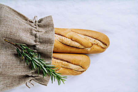 Traditional French baguette in recycled brown bag over white textile background decorated with rosemary. Sustainable package zero waste concept. Flat lay. Top Viewの写真素材