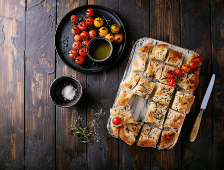 Home made focaccia flat bread cut into pieces served with baked tomatoes, olive oil and fresh rosemary, with vintage napkin and knife over a wooden background. Top Viewの写真素材