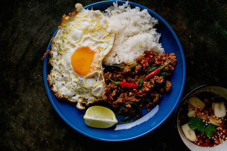 Top-down view of a traditional Thai dish, Pad Kra Pow, served on a blue plate with white rice, a sunny-side-up egg, minced pork stir-fried with basil, red chilies, and a lime wedge. A bowl of spicy dipping sauce with garlic and cilantro is on the side.の写真素材