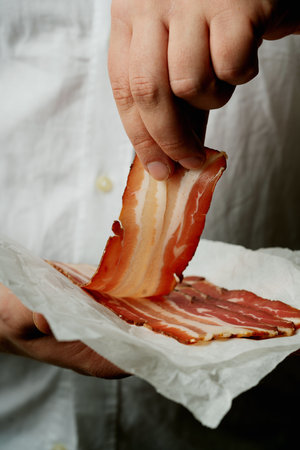 Close-up of a hand holding a thin slice of marbled bacon, lifted from a pile of slices on parchment...