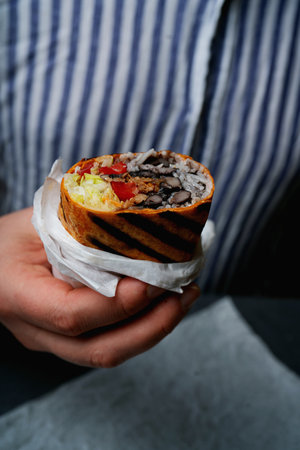 Close-up images show a person holding a rolled and grilled burrito, revealing the filling with rice, beans, tomatoes, lettuce, and meat, against a striped shirt background.の写真素材