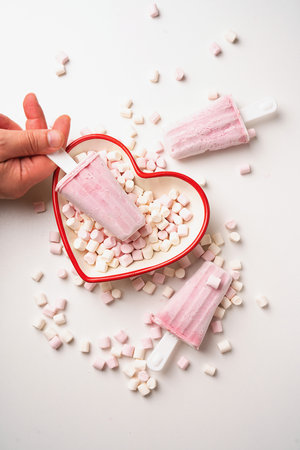 Pink ice cream popsicles arranged with marshmallow toppings,  placed in a red heart-shaped dish....