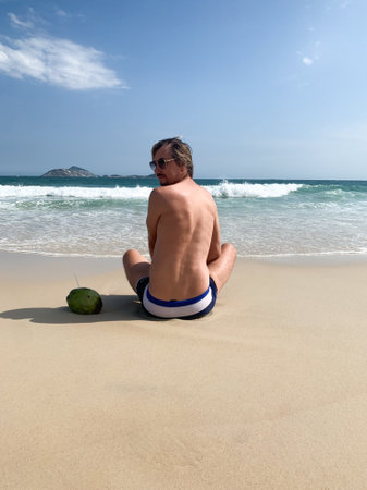 Man sitting on Copacabana Beach in Rio de Janeiro, Brazil, facing the ocean with waves and green coconut. Relaxing tropical scene under bright blue sky and sandy beach.の写真素材