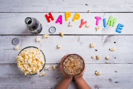 Child hand hold glass of Cola drink and Popcorn in glass bowl on white wooden table background with word happy timeの写真素材