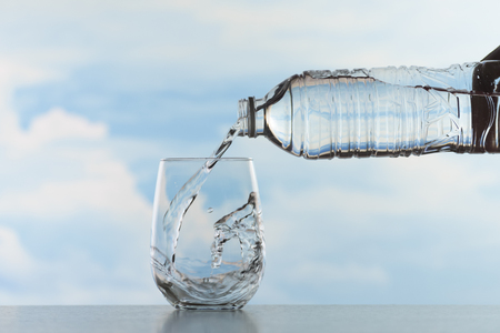 Water splashing in a transparent glass after pouring mineral water from a clear bottle with blue sky and clouds on backgroundの写真素材