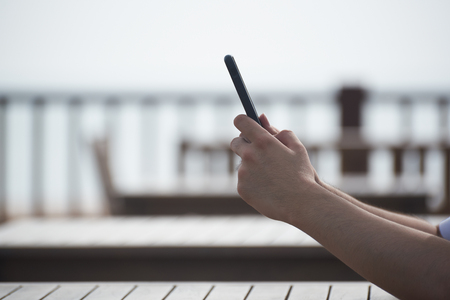 A man using both hands holding cellphone in the air and sitting near the beachの写真素材