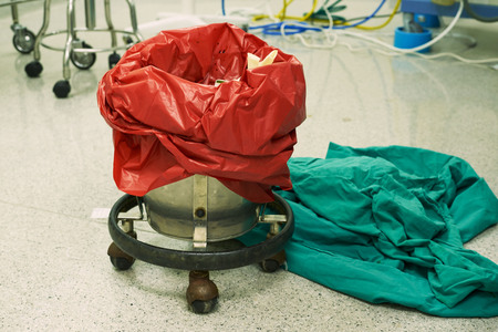 Used surgical gloves in a red garbage bin for biohazard disposal in operating room. Green clothes on the floor.の写真素材