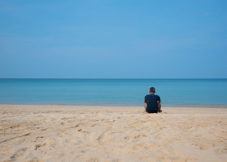 A senior Asian male sitting on the beach with the calm sea and blue sky background.の写真素材