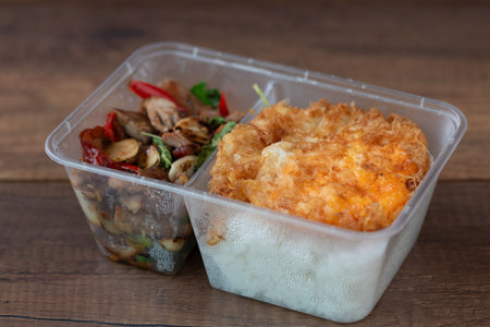 A flavorful dish of stir-fried duck with Thai basil, served over steamed rice and topped with a perfectly fried egg. Presented in a plastic takeaway container.の写真素材