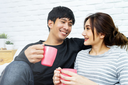 Happy Asian couple is smiling and looking each other at sofa in morning. Love and happiness concept.の写真素材