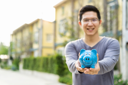 Happy asian man holding piggybank with front outdoor home background copy space on left. Smiling rich property owner loan, mortgage or installment for house. Investment in real estate business and debt conceptの写真素材