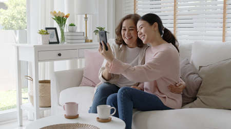 Retired healthy mom and girl two people hold phone waving hand greeting selfie look at camera on webcam app at relax comfort sofa couch living room in health care quarantine social distance isolate.の写真素材