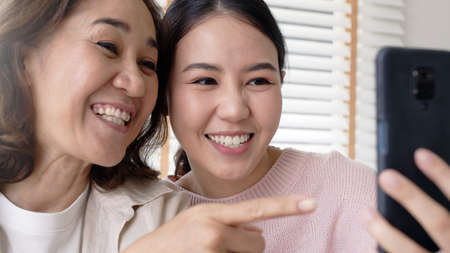 Retired healthy mom and girl two people hold phone waving hand greeting selfie look at camera on webcam app at relax comfort sofa couch living room in health care quarantine social distance isolate.の写真素材