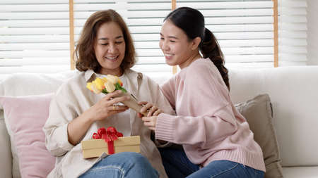 Attractive beautiful asian middle age mum sit with grown up daughter give gift box and flower in family moment celebrate mother day. Overjoy bonding cheerful kid embrace relationship with retired mom.の写真素材