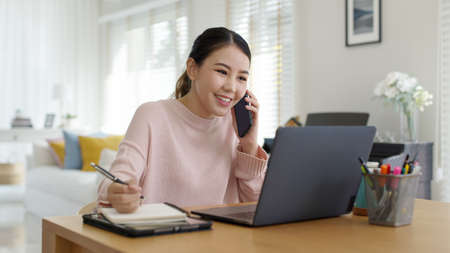 Young happy attractive beautiful asia female staff laugh talk on mobile phone relax comfort sit work on desk table at home. Remotely work on distance job telesale or secretary career for business.の写真素材