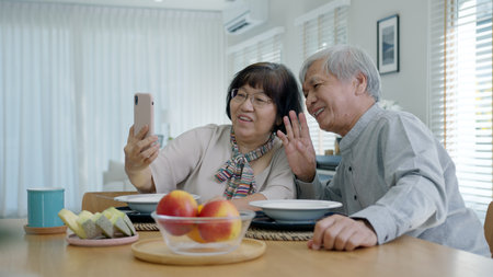 Young senior asia citizen couple hold mobile phone selfie videocall online to family at home apartment morning on dining table in older people with digital technology virtual talk conference concept.の写真素材