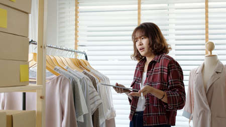 Young attractive beautiful hipster asia female fashion store owner use digital tablet check stock clothes or package box on rail rack at home office small SME business entrepreneur e-commerce concept.の写真素材