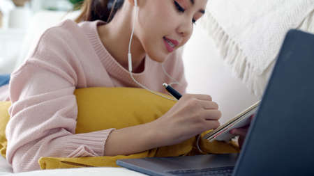 Cozy relax young attractive asian female employee wear earphone listen to digital online course development program in quarantine social distance at home reskill upskill school class for career path.の写真素材