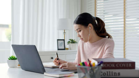 Candid young attractive asian female employee wear earphone listen to digital online course development program in quarantine social distance at home reskill upskill school class for career path.の写真素材